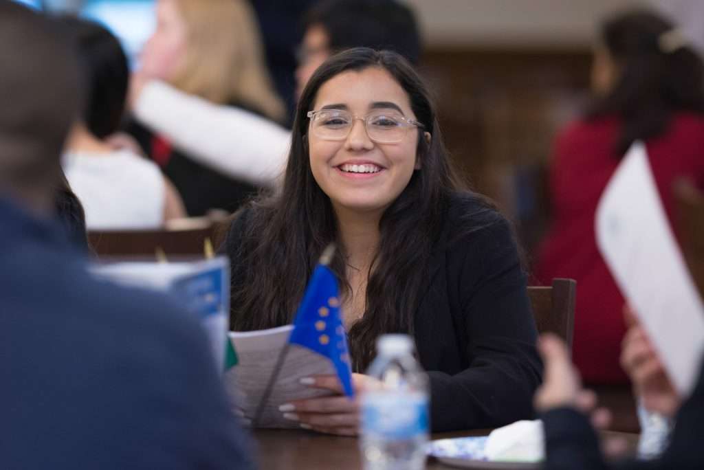A female student smiling brightly while holding a paper, participating in discussions at a formal event with peers.