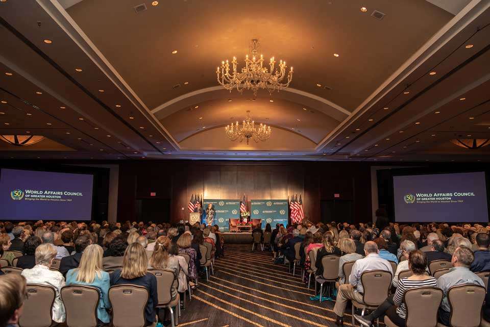 Large audience attending a World Affairs Council event in a grand ballroom with chandeliers, featuring two screens displaying the council’s 50th anniversary logo.
