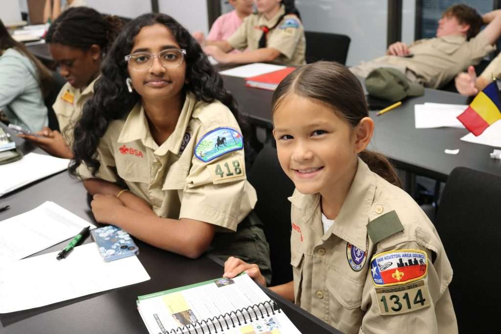 Two girl scouts, one with glasses and one smiling, sit next to each other at a table, with other scouts working in the background.