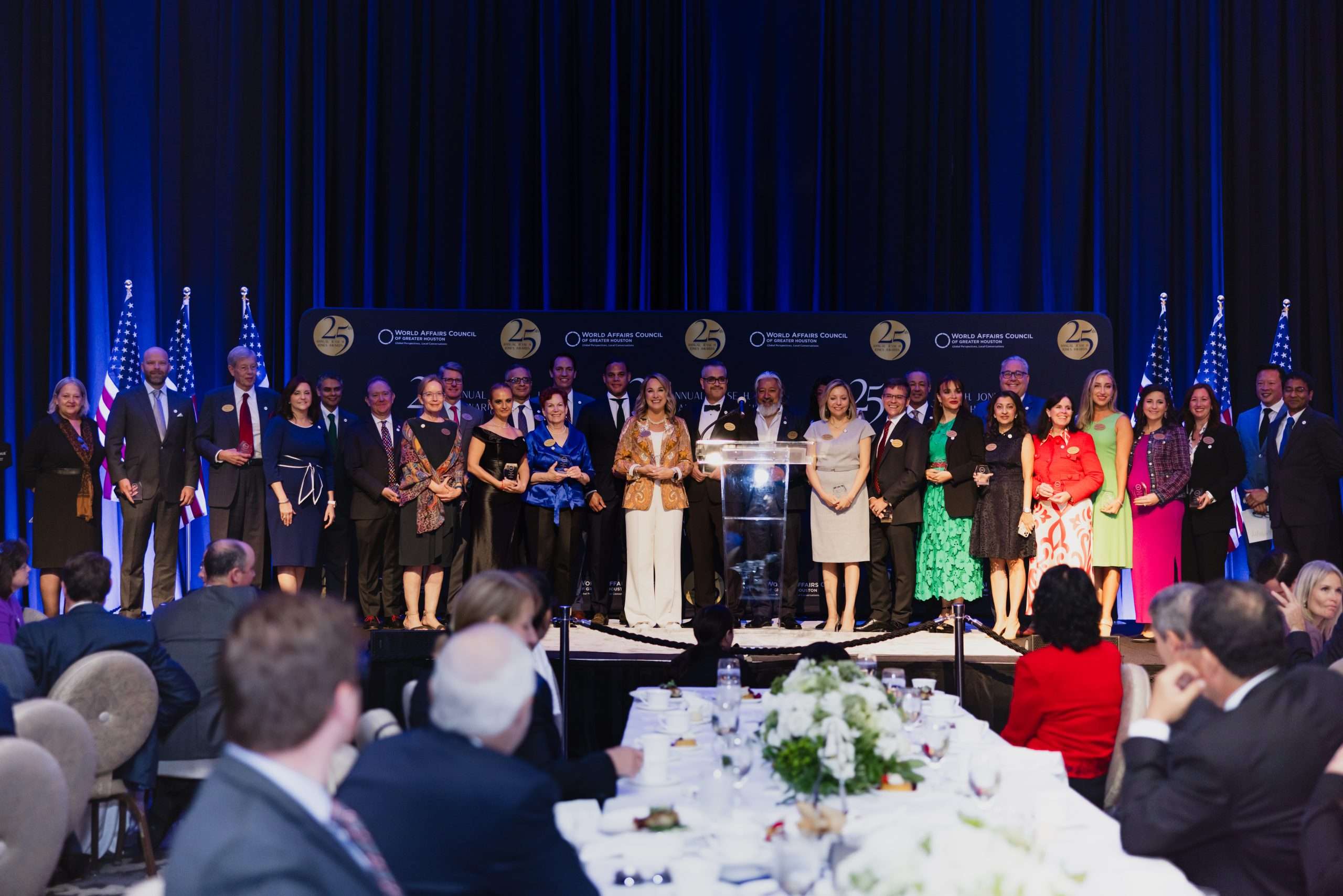 Large group of award recipients and speakers stand on stage at an event hosted by the World Affairs Council of Greater Houston, with blue and gold decor and American flags in the background.
