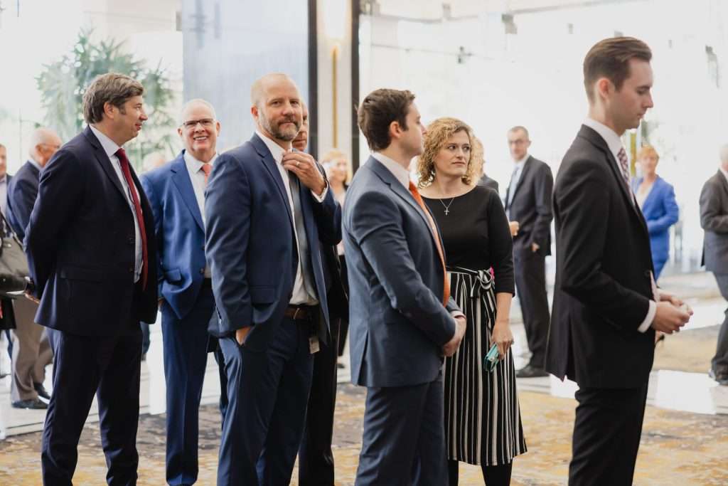 People in business attire standing in a lobby area, some engaging in conversation, with a bright and modern background.