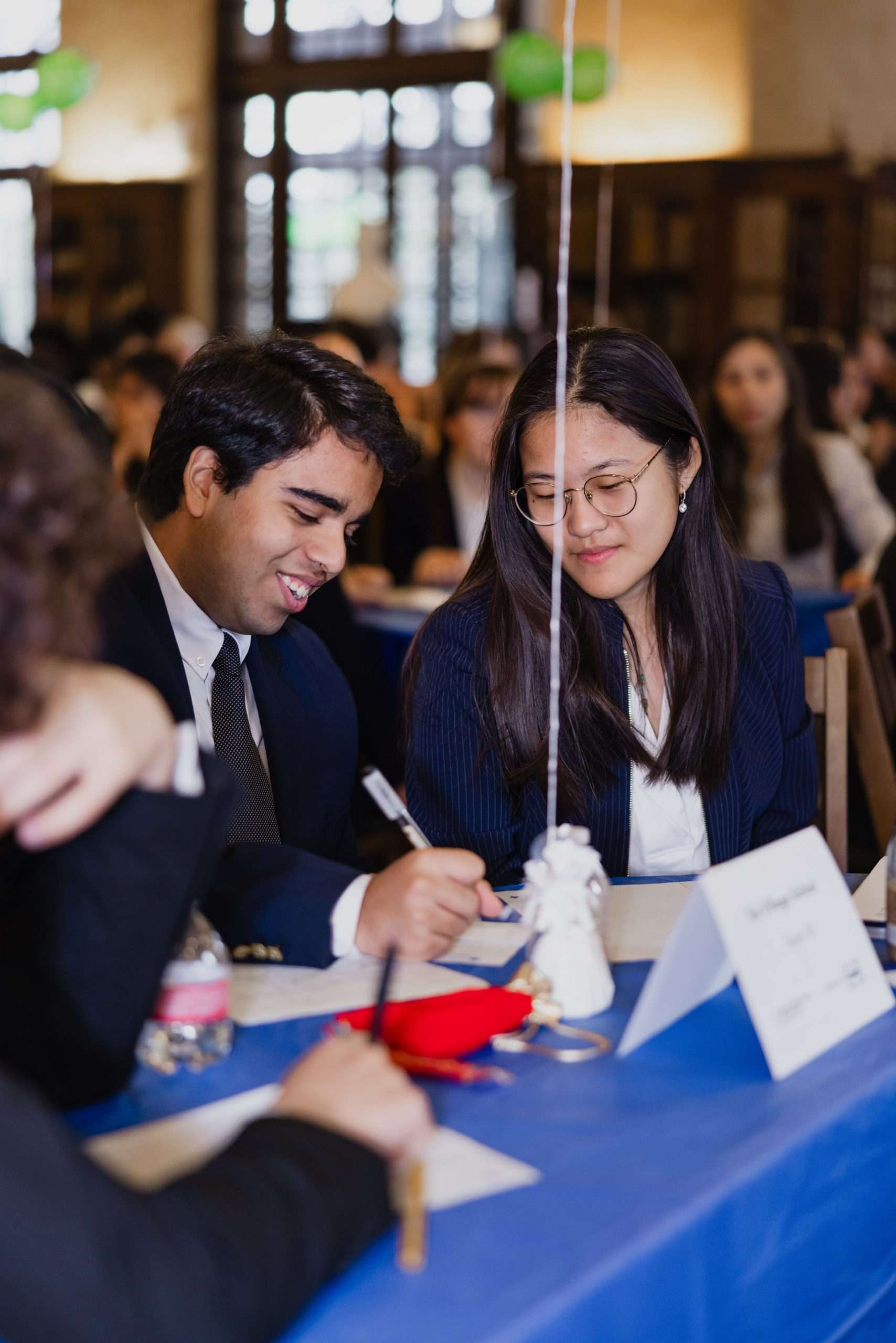 Two people at a table smiling and writing, one with glasses and dark hair.