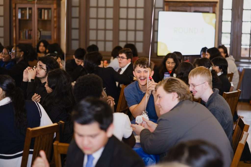 A lively group of students sitting at round tables in a large room, participating in an activity. One student in the middle holds up a balloon with a joyful expression, while others are smiling and engaged in conversation.