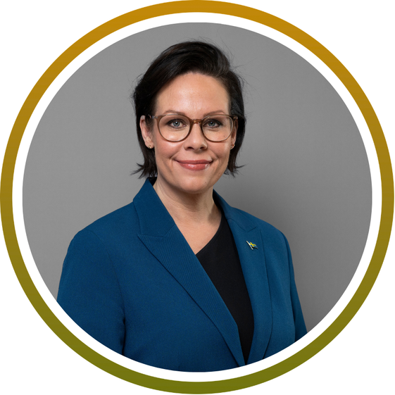 A person with short dark hair and glasses wearing a blue blazer over a black top, smiling, posed in front of a gray background with a circular gold and white border around the image.