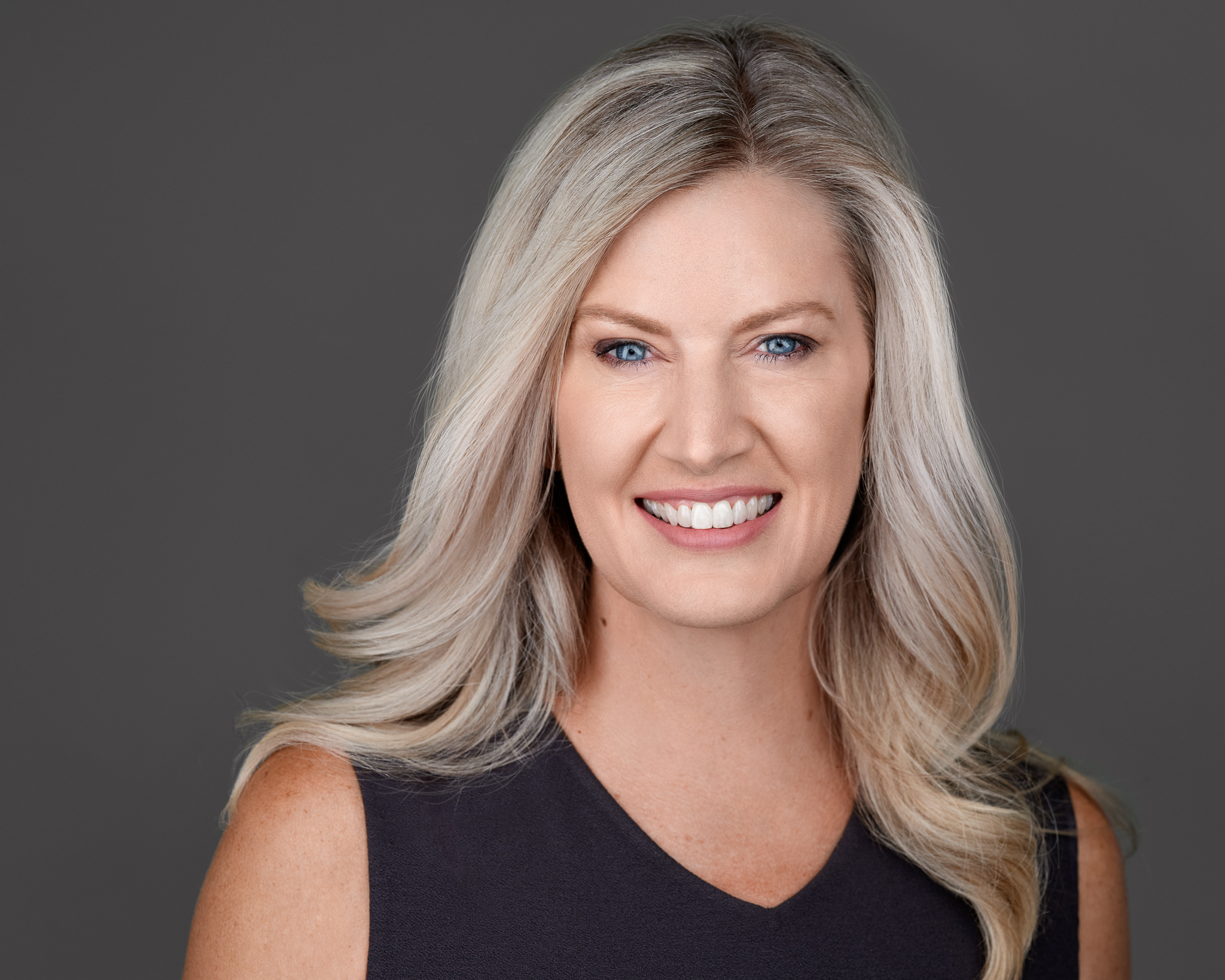 Smiling woman with long, wavy blonde hair wearing a sleeveless black top, posing in front of a plain grey background.