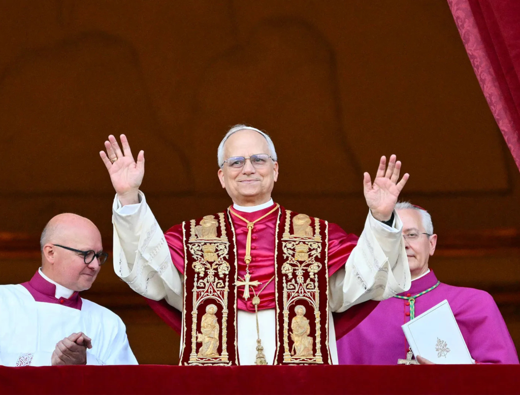 A man in ornate religious robes raises both hands while standing on a balcony between two other men in clerical attire, against a red and gold backdrop.