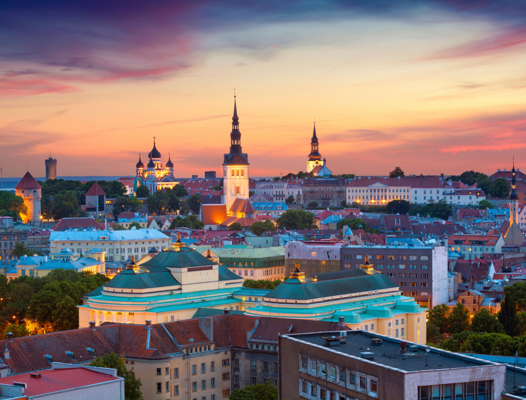 Colorful cityscape of Tallinn, Estonia at sunset, with historic churches, domes, and rooftops illuminated by the warm evening sky, framed by lush green trees in the foreground.