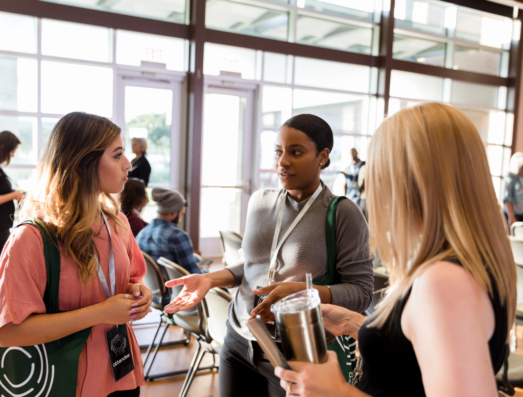 Three women stand talking in a bright, modern room with large windows. They wear conference badges and tote bags, and one woman holds a notebook while another has a tumbler. Other people sit in the background.