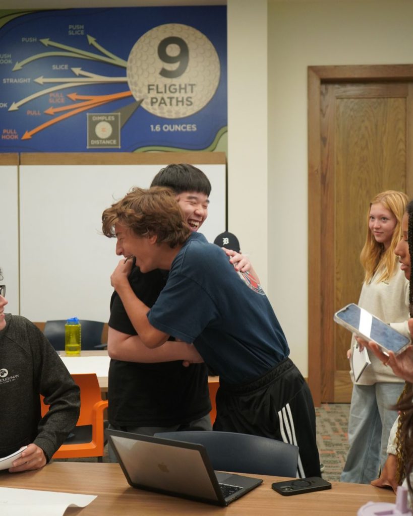 Two teenagers smile and hug each other in a classroom, surrounded by others. A Flight Paths diagram is on the wall behind them, and laptops and papers are on the table in front.