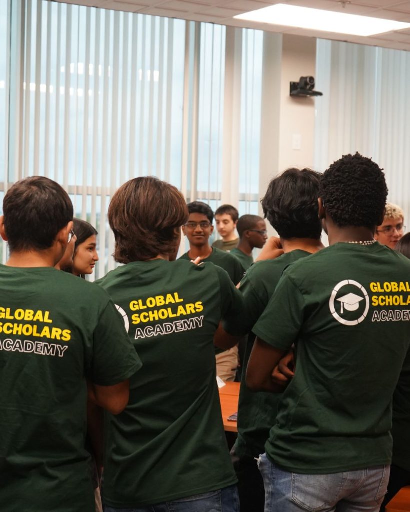 A group of students wearing matching green Global Scholars Academy t-shirts stand together and talk in a classroom with large windows and vertical blinds.