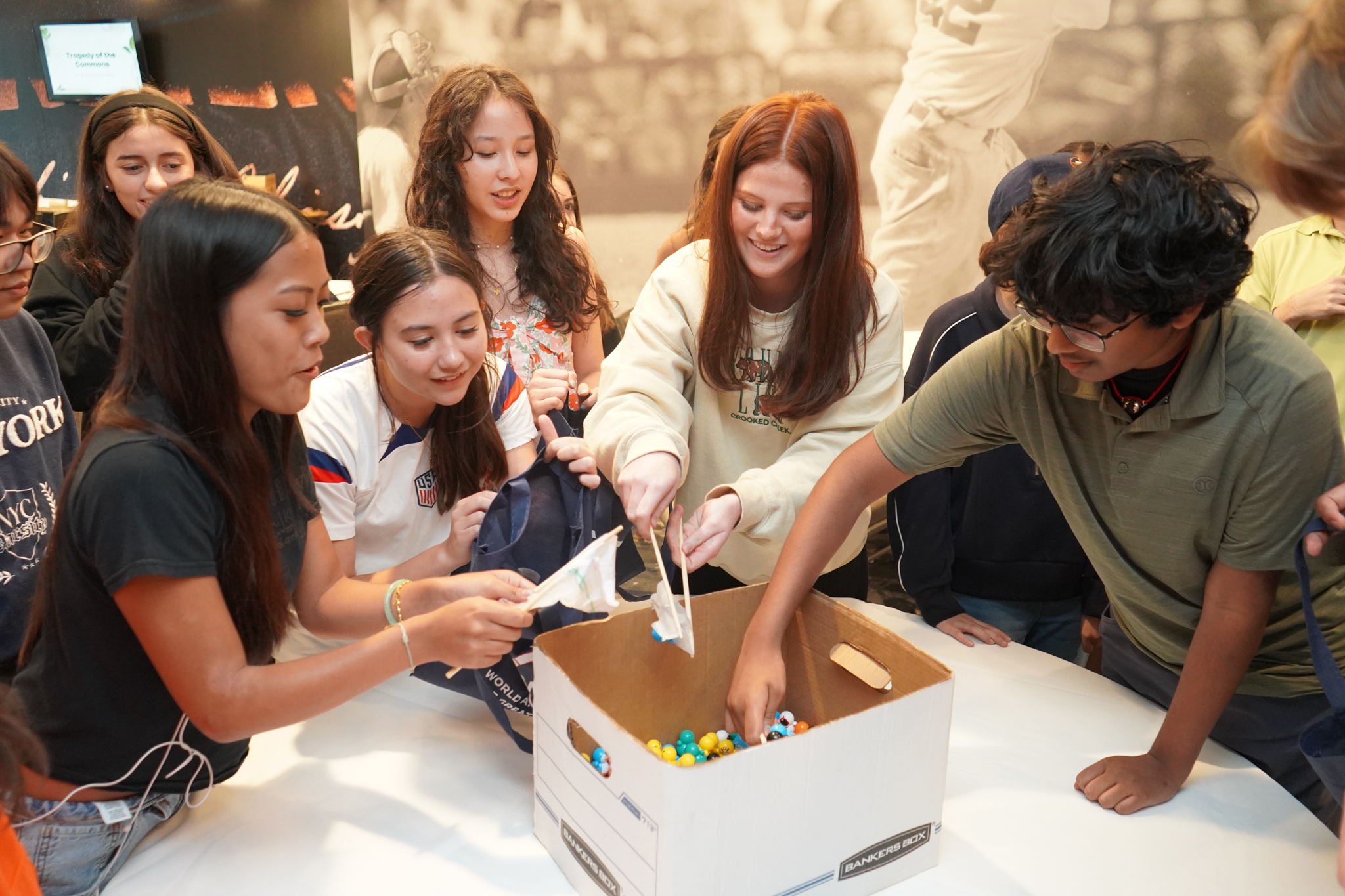 A group of smiling young people gather around a table, reaching into a large cardboard box filled with colorful plastic balls, appearing excited and engaged.