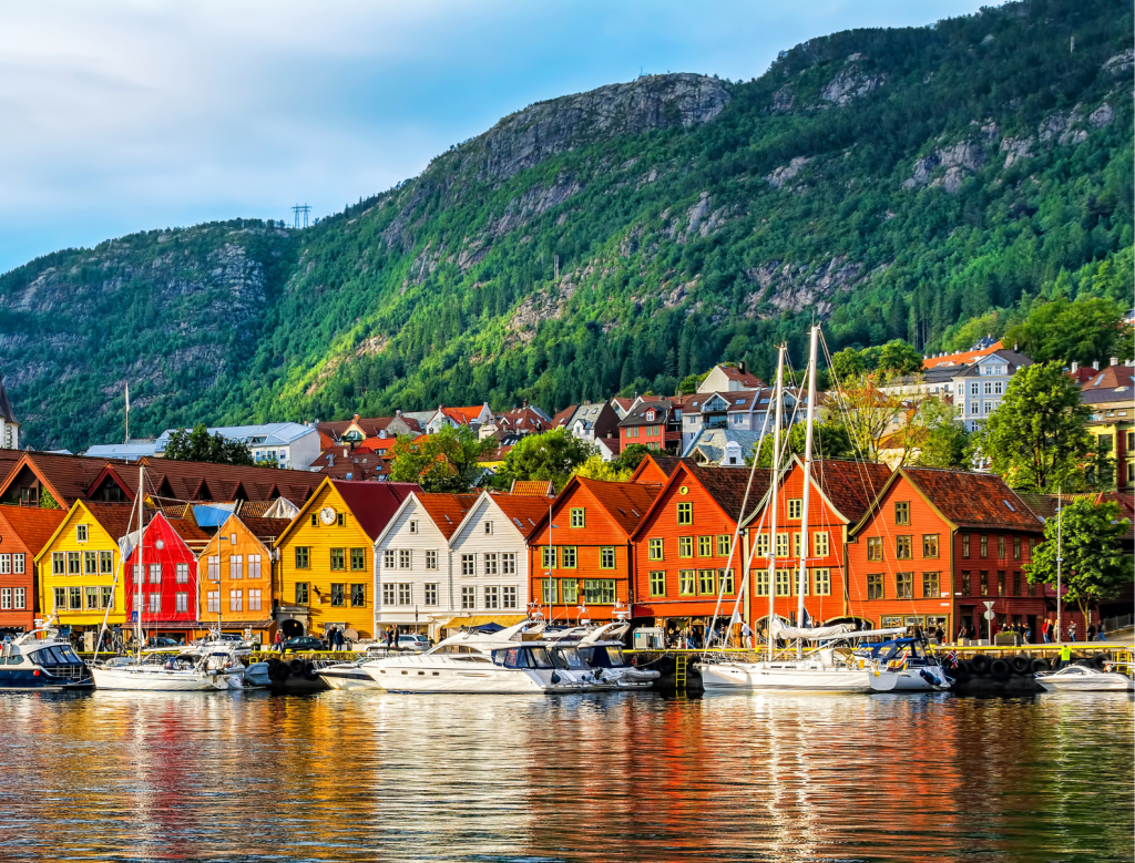Colorful wooden houses line the waterfront in front of green, forested hills; several boats are docked along the calm water reflecting the vibrant buildings.